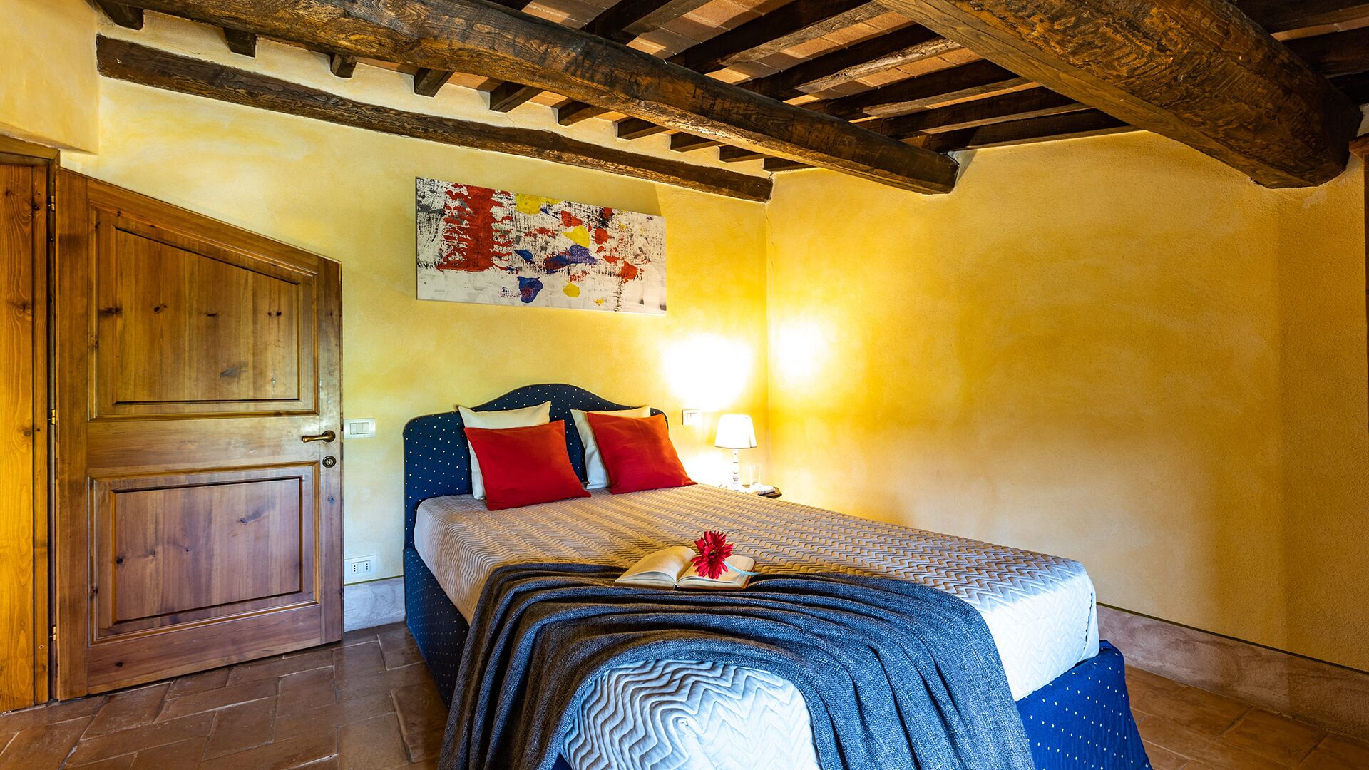 Une chambre avec une porte en bois, un lit recouvert d’une literie à motifs, un tableau accroché au mur et un sol carrelé.