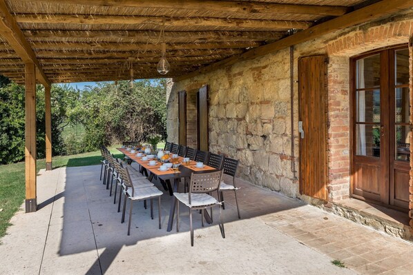 Une terrasse extérieure avec une longue table dressée pour les repas, entourée de murs en pierre et d’une pergola en bois.