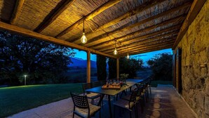 An outdoor patio with a wooden table and chairs, surrounded by stone walls, under a thatched roof with hanging light fixtures. - Il Poderuccio 17, Emma Villas (Cetona (Siena))