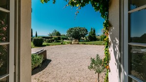 A view through an open doorway to a courtyard with a gravel path, a large tree, and a well-maintained garden. - Charming farmhouse with private heated pool and amazing mountain views. (Mont-De-Marrast)
