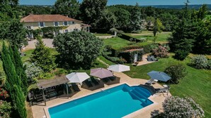 Une piscine entourée de chaises longues et de parasols, elle-même bordée d’un jardin bien entretenu, avec une maison en arrière-plan.