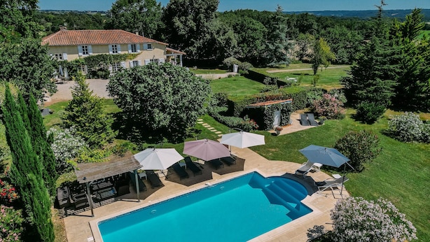 A swimming pool with lounge chairs and umbrellas, surrounded by a well-maintained garden and a house in the background. - Charming farmhouse with private heated pool and amazing mountain views. (Mont-De-Marrast)