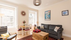 A living room with a grey sofa, a wooden chest, a red patterned rug, and a white chair. - Luxury Thistle New Town 1 Bed Apt Super King (Edinburgh)
