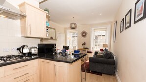 A compact kitchen with wooden cabinets, a black countertop, and a dining area with a grey sofa and a small table. - Luxury Thistle New Town 1 Bed Apt Super King (Edinburgh)