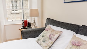 A neatly made bed with floral pillows, a bedside table with a lamp, and a window with a view of a stone building. - Luxury Thistle New Town 1 Bed Apt Super King (Edinburgh)