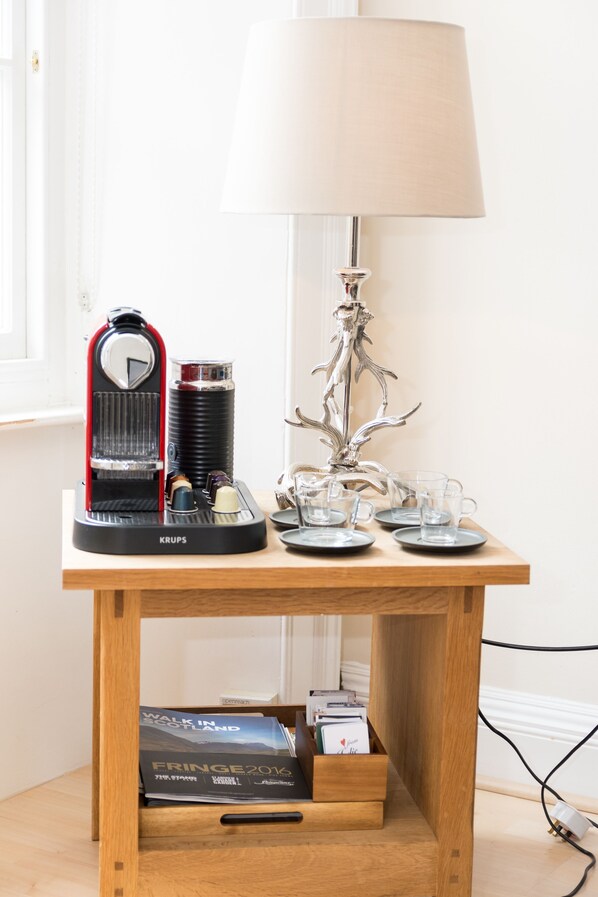 A wooden table with a Krups coffee maker, a lamp, and a few cups and saucers. There is a drawer under the table and a magazine titled "Walk in Scotland Fringe 2016" on the floor.
