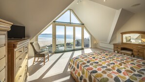 Ein Schlafzimmer mit einem großen Erkerfenster, einem Bett mit farbenfroher Musterdecke, einer Holzkommode, einem Stuhl und einem Fernseher auf einer Kommode.
