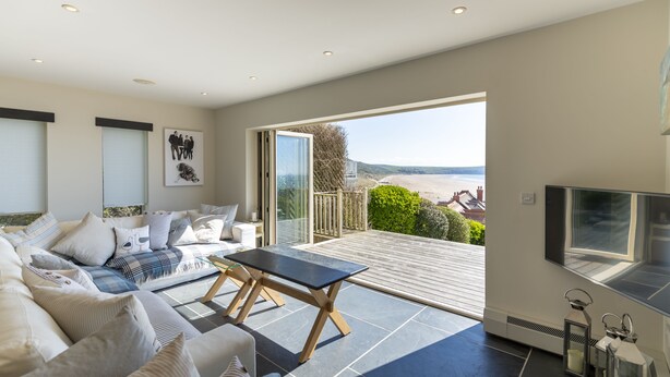 Ein modernes Wohnzimmer mit einer Couch, einem Couchtisch und einem Fernseher. Ein großes Fenster bietet Blick auf den Strand.
