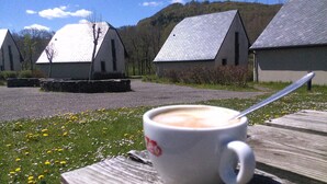 Une tasse de café blanche est posée sur une table en bois, avec une cuillère à côté. L’ensemble est mis en valeur par un décor de bâtiments traditionnels aux toits inclinés, entourés d’une zone herbeuse parsemée de fleurs jaunes.