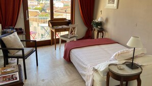 Habitación de hotel con una cama, un escritorio con silla, una mesita con lámpara y vistas a la ciudad a través de amplias ventanas.