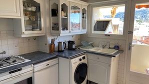 Una cocina compacta con muebles blancos, estufa a gas, lavadora y ventana con vista a edificios.
