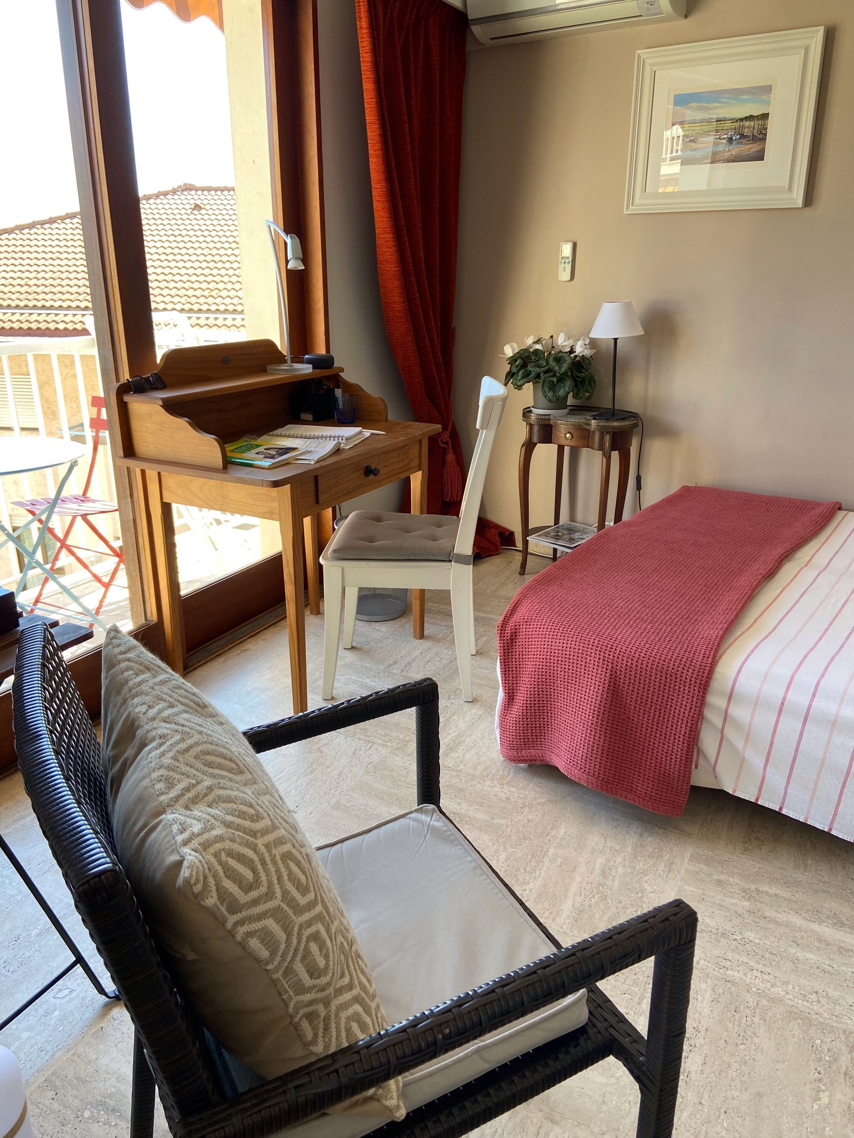 A room with a bed, a desk with a chair, a wicker chair, a small table with a lamp, and a window with curtains.