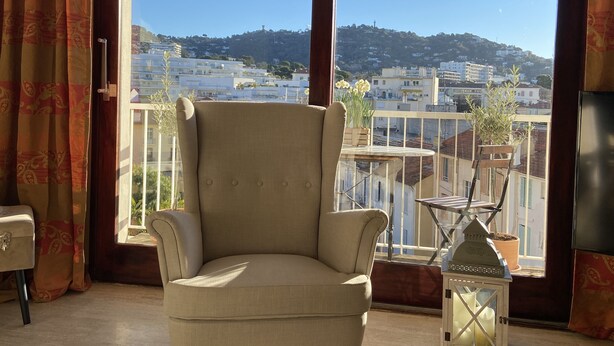 Una habitación con un sillón color beige, una mesita con una planta en maceta y vistas a un paisaje urbano a través de amplios ventanales.