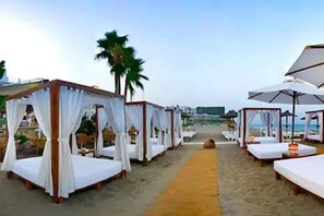 Un hôtel situé en bord de mer, doté de plusieurs lits à baldaquin blancs disposés le long d’une promenade de sable.