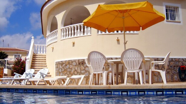 Un espace aménagé au bord de la piscine, avec des chaises longues et un parasol, un mur en pierre et un bâtiment doté d’un balcon.