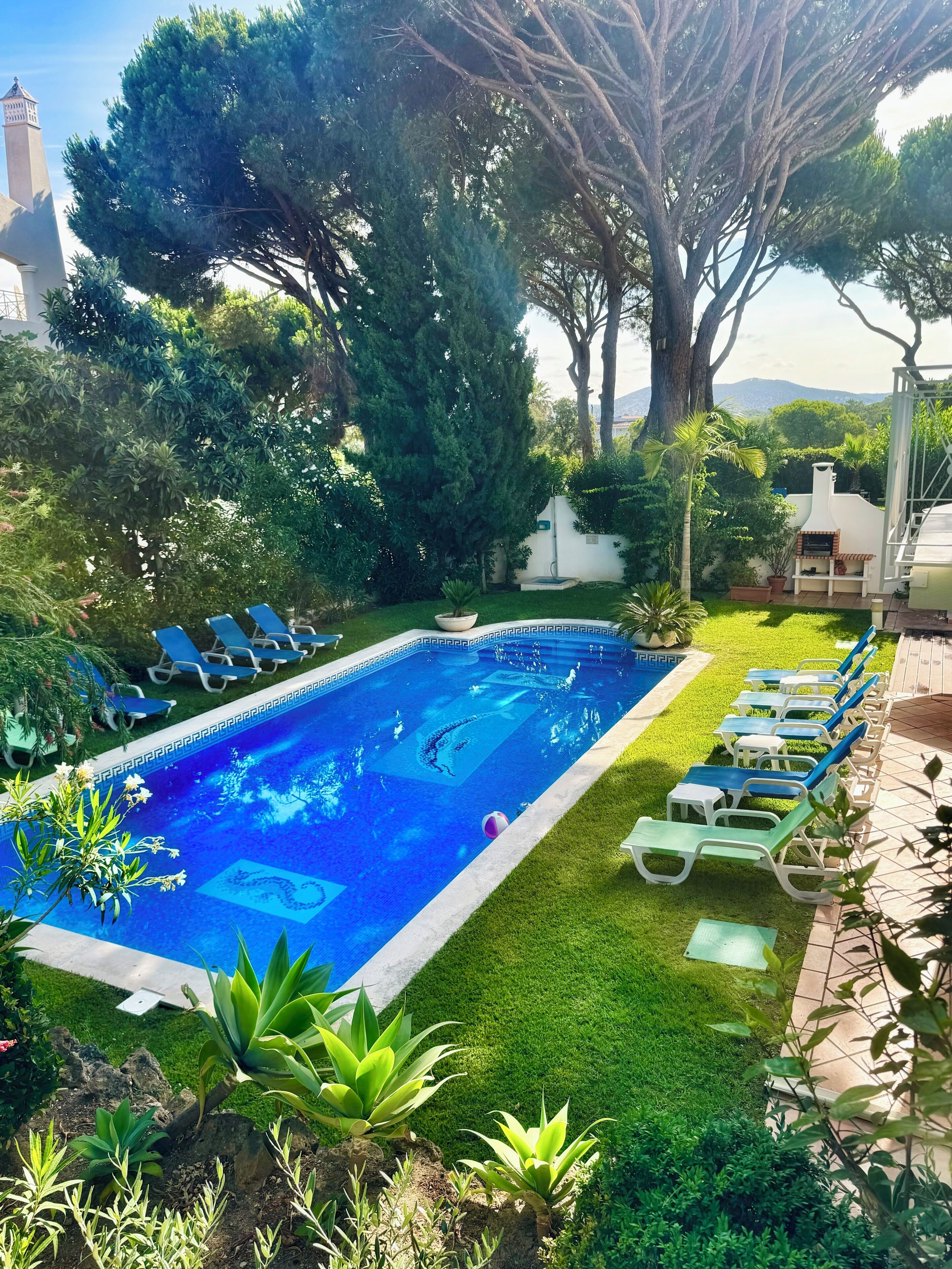 A swimming pool with lounge chairs, surrounded by greenery and a view of mountains.