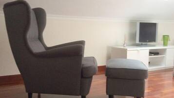 Une pièce avec un parquet en bois foncé, un fauteuil gris, un pouf assorti, une armoire blanche et un téléviseur posé sur un meuble.