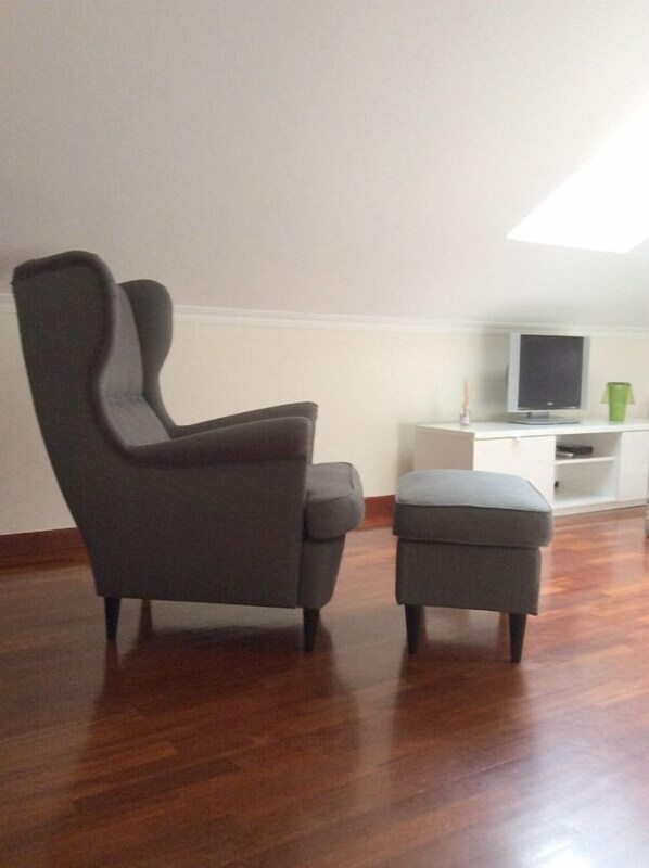Una habitación con piso de madera oscura, un sillón gris, un otoman a juego, un armario blanco y un televisor sobre un soporte.