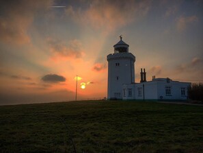 A lighthouse with a cylindrical tower and a smaller building attached to it, set against a sunset sky.