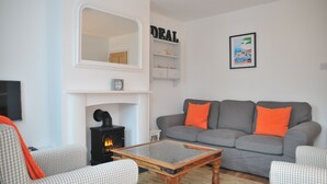A living room with a fireplace, a grey sofa with orange cushions, a checkered armchair, and a coffee table. - Goodwin Sands Cottage - 50 meters from the beach  (Deal)