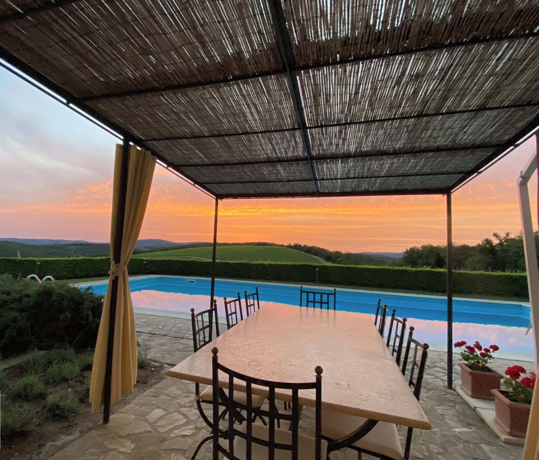 A covered outdoor dining area with a pool view, stone flooring, and a table with chairs.