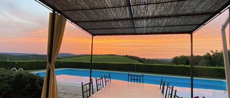 A covered outdoor dining area with a pool view, stone flooring, and a table with chairs.
