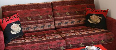 A red sofa with patterned cushions and two pillows with 'MOCHA' text. A tray with a teapot, cups, and saucers is on the table in front.
