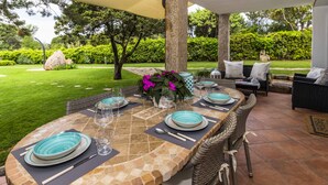 An outdoor dining area with a long table set for six, surrounded by wicker chairs, a cushioned sofa, and a large umbrella.
