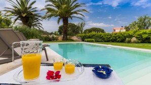 A poolside setting with a glass pitcher and two glasses of orange juice, a blue bowl, and a red flower on a tray.