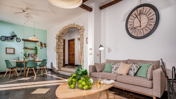 Ein Wohnzimmer mit Couch, Holz-Couchtisch, gemusterten Kissen, einer großen Wanduhr und einem Essbereich mit Stühlen und Tisch.