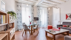 A modern living room with a glass dining table, grey chairs, a grey sofa, and a red ottoman. There is a bookshelf with books and a potted plant. - Grotta Pinta 3 | Spacious and elegant just few steps from Piazza Navona (Roma)