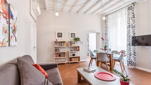 A modern living room with a grey sofa, a dining table with chairs, and a TV mounted on the wall. There are bookshelves with books and decorative items, and a potted plant on the table. - Grotta Pinta 3 | Spacious and elegant just few steps from Piazza Navona (Roma)
