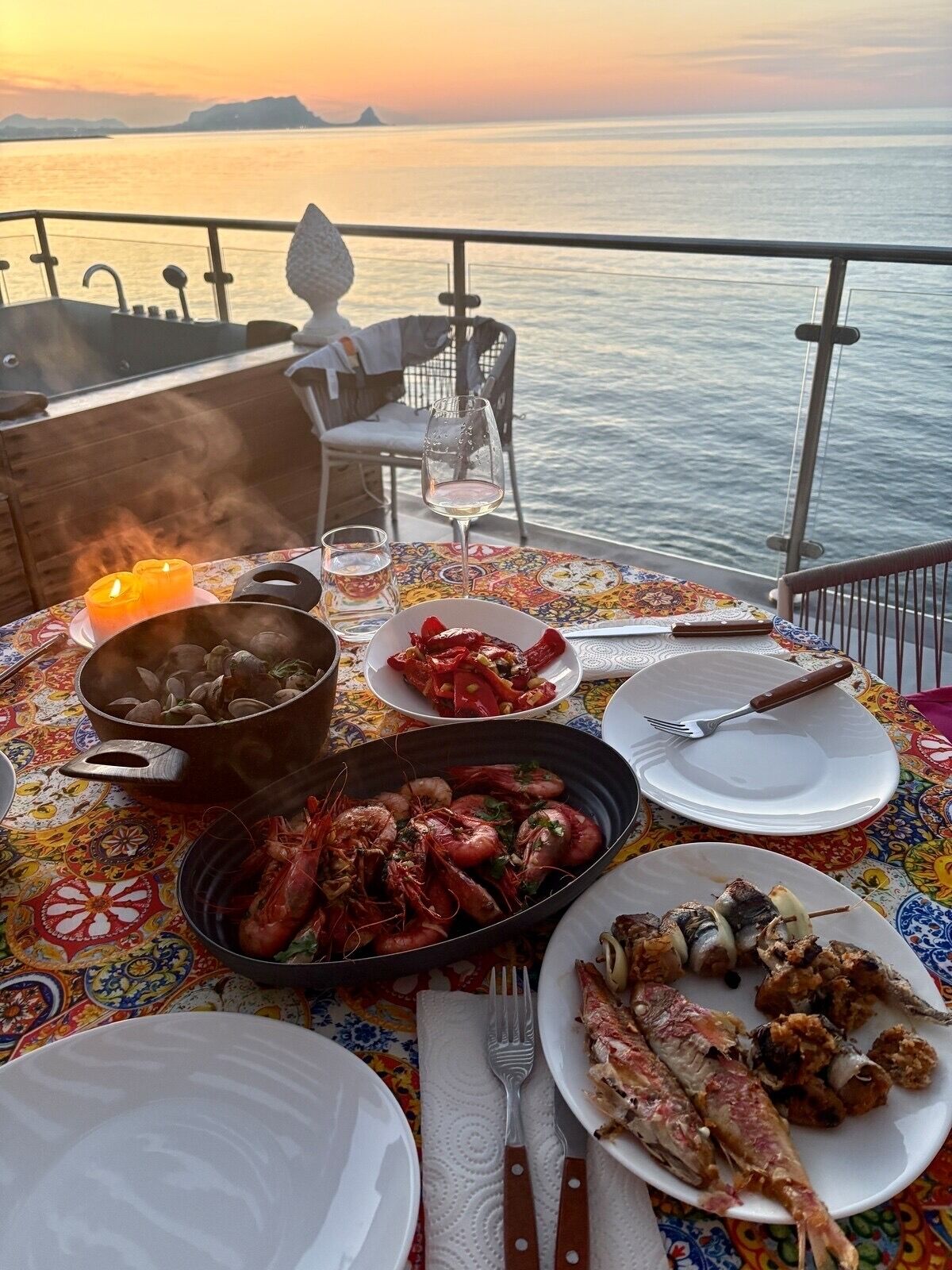 Une table dressée avec des plats de fruits de mer, un verre de vin et une bougie, donnant sur une étendue d’eau au coucher du soleil.