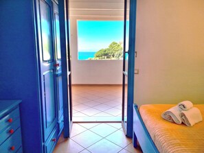 A room with a bed, a window with a view of the sea, and a blue wardrobe.