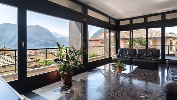 A spacious living room with large windows, a black leather sofa, a glass coffee table, and a potted plant. - Terrace on the lake,private garage (Lugano)