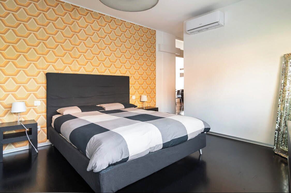 Ein modernes Hotelzimmer mit Wand im geometrischen Muster, einem großen Bett mit schwarzem Kopfteil und einem kleinen Nachttisch mit Lampe.