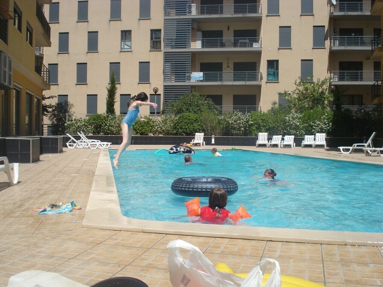 Una piscina con gente nadando y jugando, rodeada de sillas de descanso y un edificio al fondo.