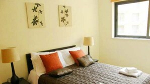 Ein ordentlich bezogenes Bett mit brauner Tagesdecke und Kissen, zwei Wandlampen und ein Bild an der Wand.