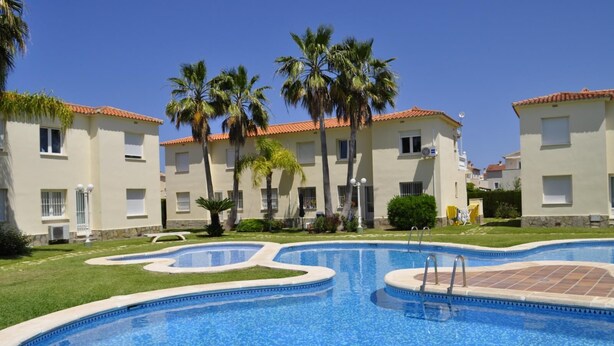A swimming pool with a tiled deck, surrounded by palm trees and buildings with red-tiled roofs.