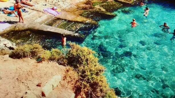 Une zone côtière caractérisée par une eau bleue et limpide, un terrain rocheux et des personnes qui se baignent.