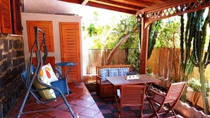 Eine Terrasse im Freien mit einem Holztisch und Stühlen, einem blauen Klappstuhl und einer gepolsterten Bank.