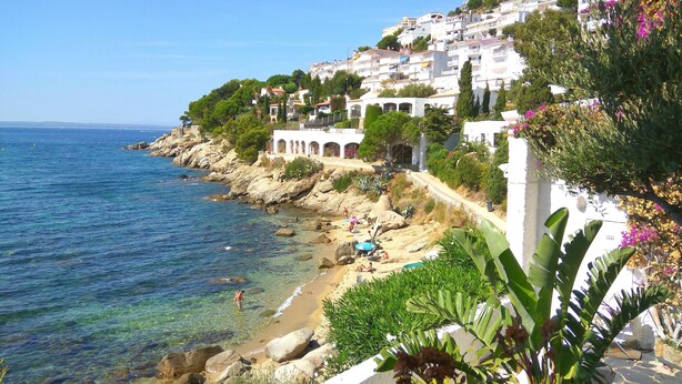 Eine Küstenstadt mit Gebäuden, die sich an einen Hügel schmiegen und auf das Meer blicken, mit einem Sandstrand und klarem, blauem Wasser.