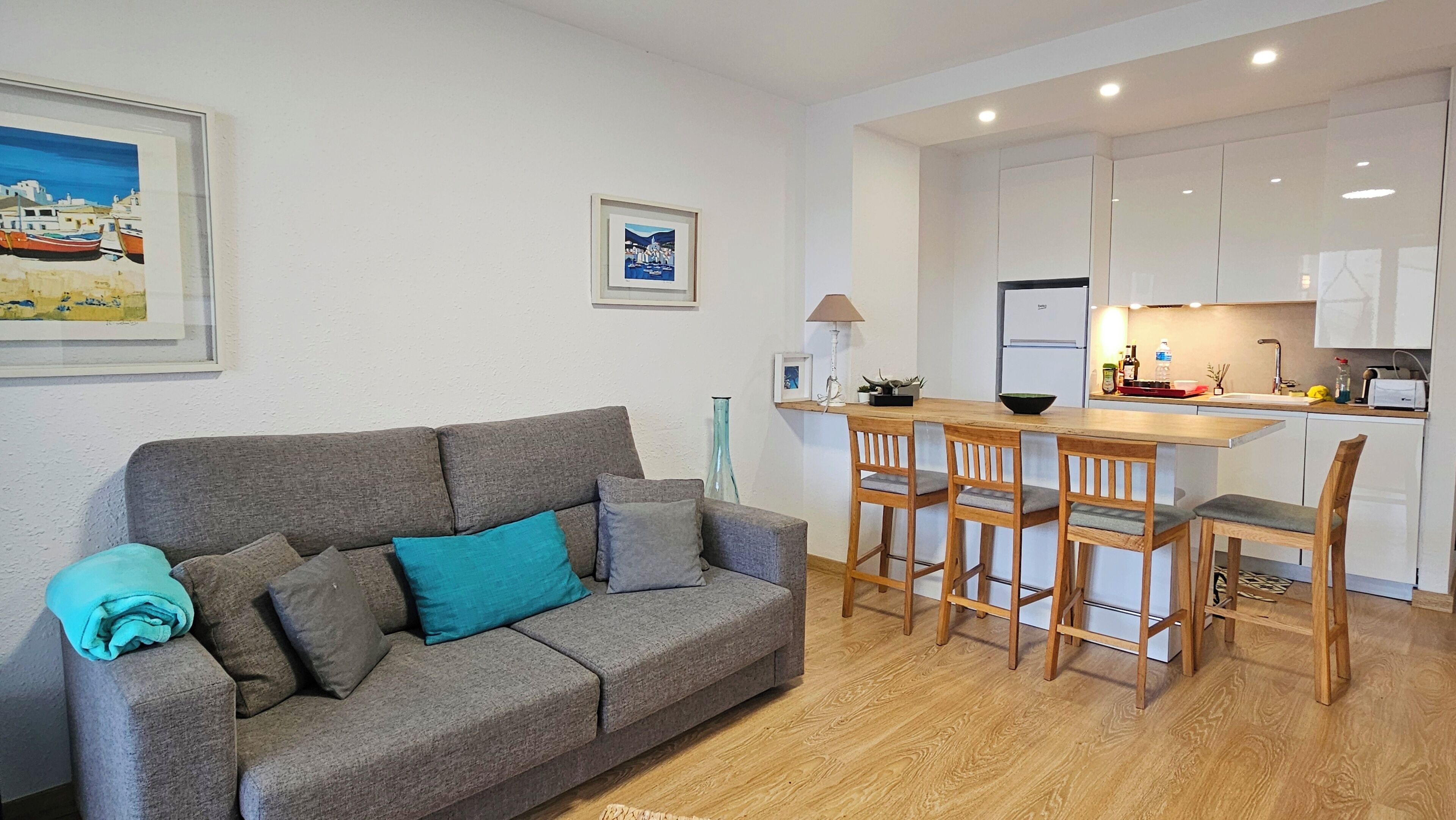 Una sala moderna con un sofá gris, una mesa de comedor de madera con sillas, una zona de cocina con armarios blancos y un cuadro en la pared.