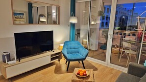Ein modernes Wohnzimmer mit Fernseher, einem blauen Sessel, einem kleinen Tisch mit Obst und einem Balkon mit Blick auf Gebäude.
