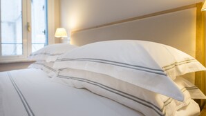 Un lit bien fait, avec une literie blanche à rayures grises, une lampe de chevet et une fenêtre avec des stores.