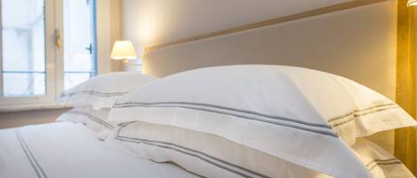 Una cama bien hecha con ropa blanca a rayas grises, una lámpara de noche y una ventana con persianas.
