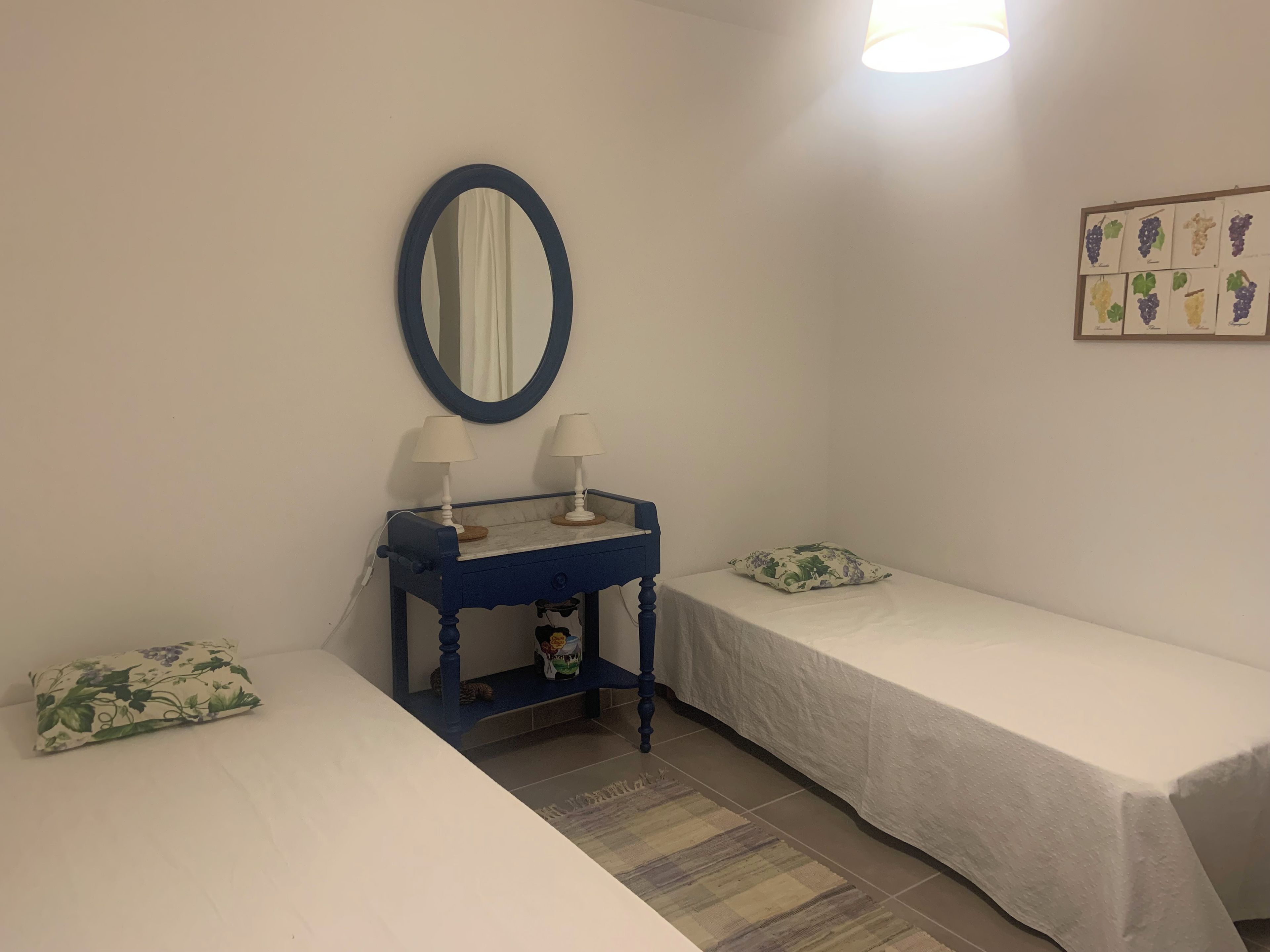 Ein Zimmer mit zwei Einzelbetten, einem kleinen Tisch mit zwei Lampen, einem Spiegel und einem an der Wand hängenden Bild von Trauben.