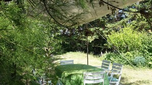 An outdoor patio with a round table and white chairs, surrounded by lush greenery and a large umbrella providing shade.