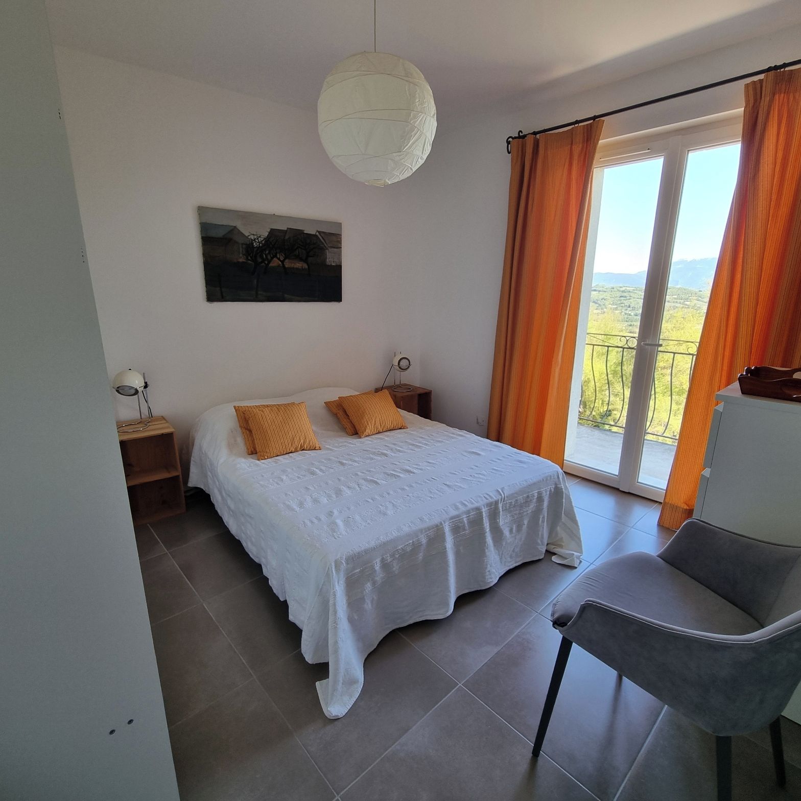 A bedroom with a bed, a chair, a bedside table, a lamp, a painting, and a view through a window.