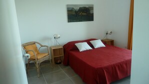 A bedroom with a red bedspread, two white pillows, a wicker chair, a nightstand with a lamp, and a painting on the wall.
